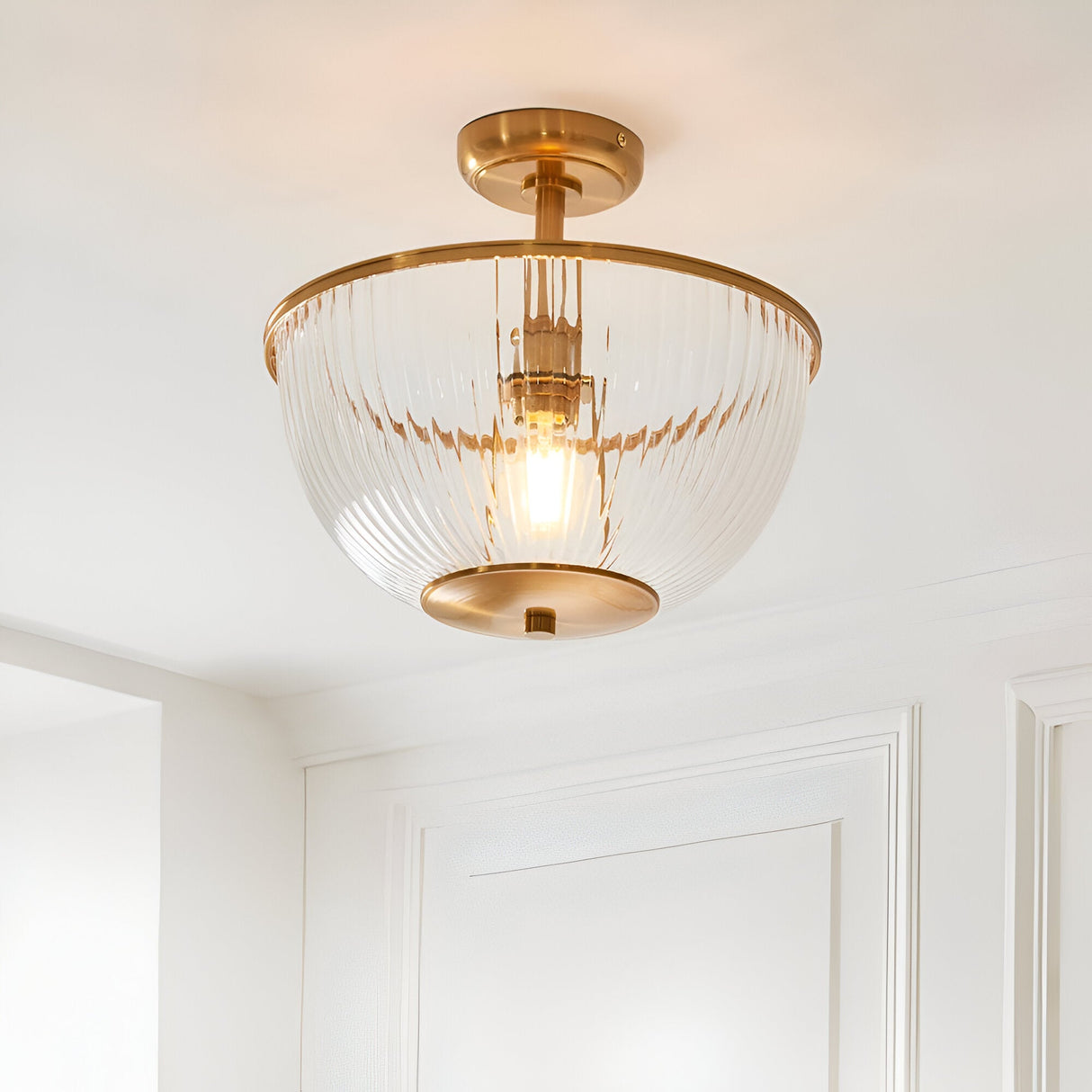 Endon Langton flush uplighter in brass with a ribbed glass bowl diffuser and single E27 lamp, mounted to a white ceiling above a panelled wall.