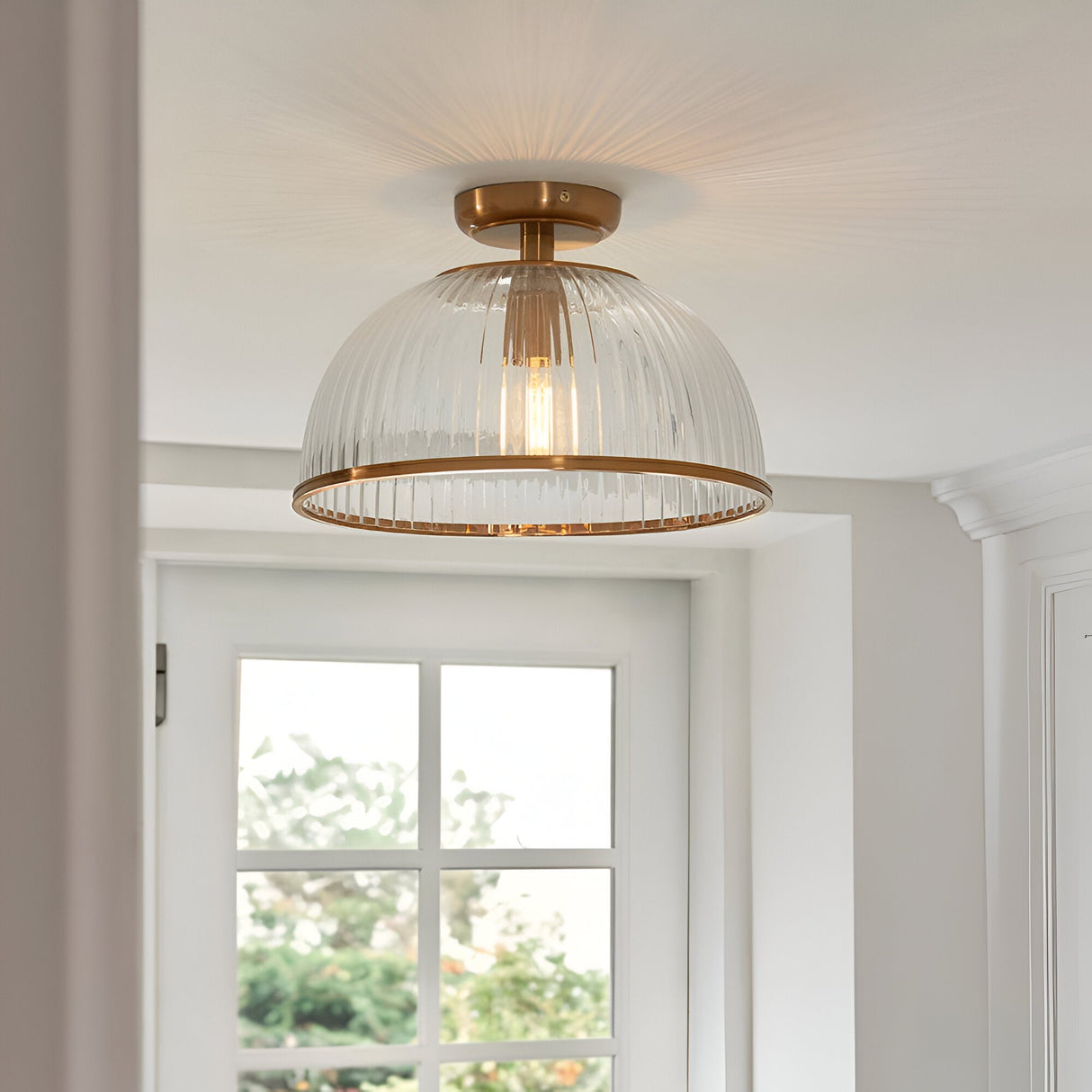 Langton Flush Downlight in brass with ribbed glass dome shade, shown mounted close to a white ceiling in a hallway near a window.