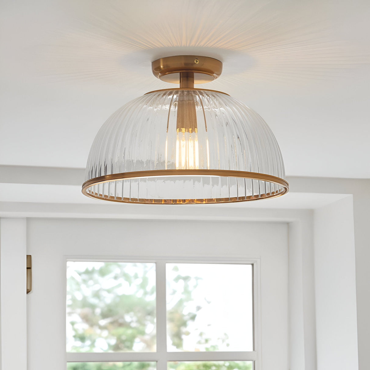 Langton Flush Downlight in brass with ribbed glass dome shade and exposed E27 bulb, fitted to a white ceiling above a bright room with window.