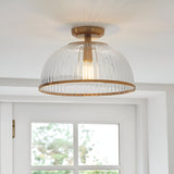 Langton Flush Downlight in brass with ribbed glass dome shade and exposed E27 bulb, fitted to a white ceiling above a bright room with window.