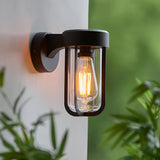 Caldwell Wall Light in matt black die-cast aluminium with clear glass shade and visible E27 bulb, mounted outdoors near green foliage.