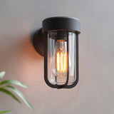 Caldwell Wall Light in matt black die-cast aluminium with clear glass shade and exposed E27 bulb, mounted on a light grey wall.