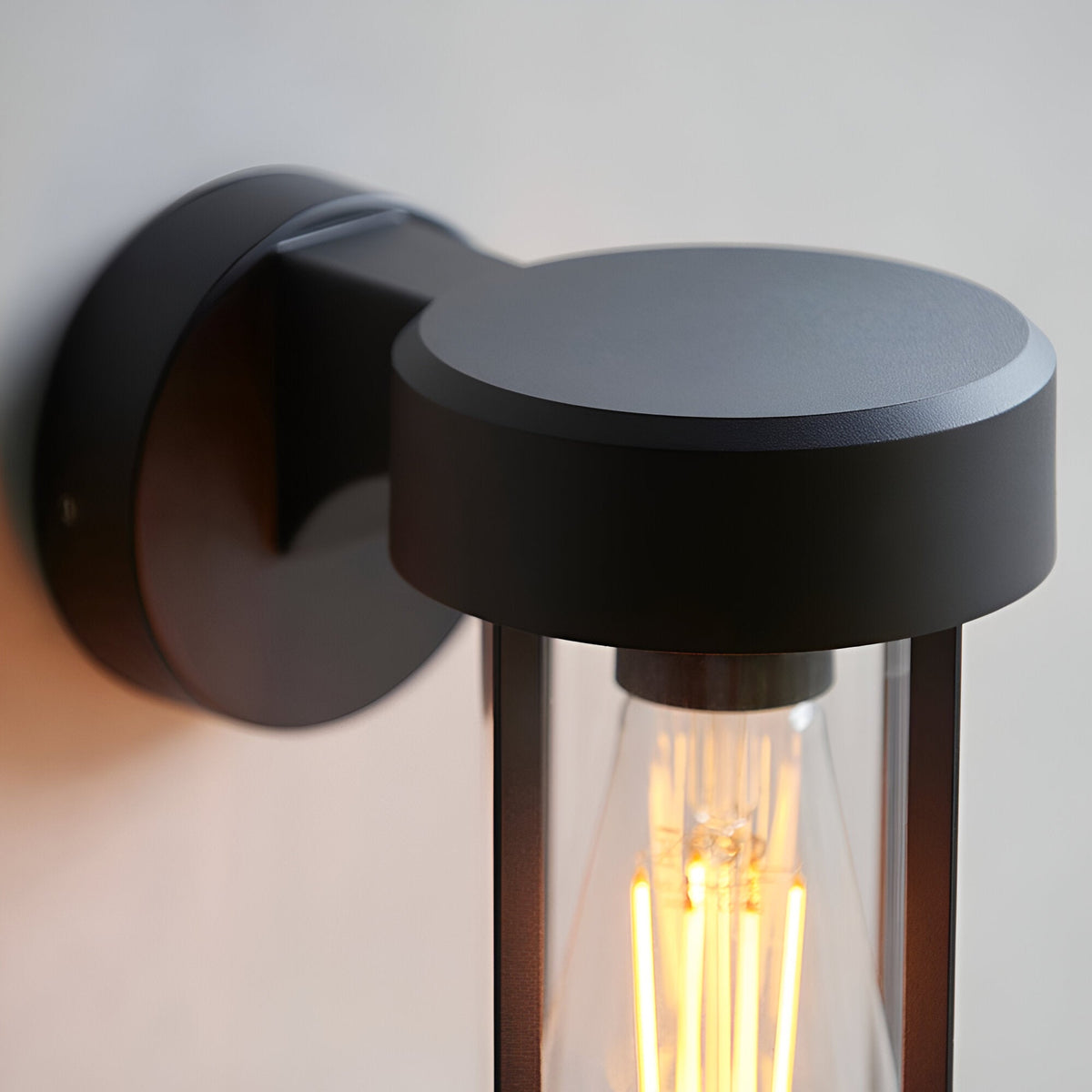 Caldwell Wall Light in matt black die-cast aluminium with a round backplate and cylindrical glass shade housing an E27 bulb.