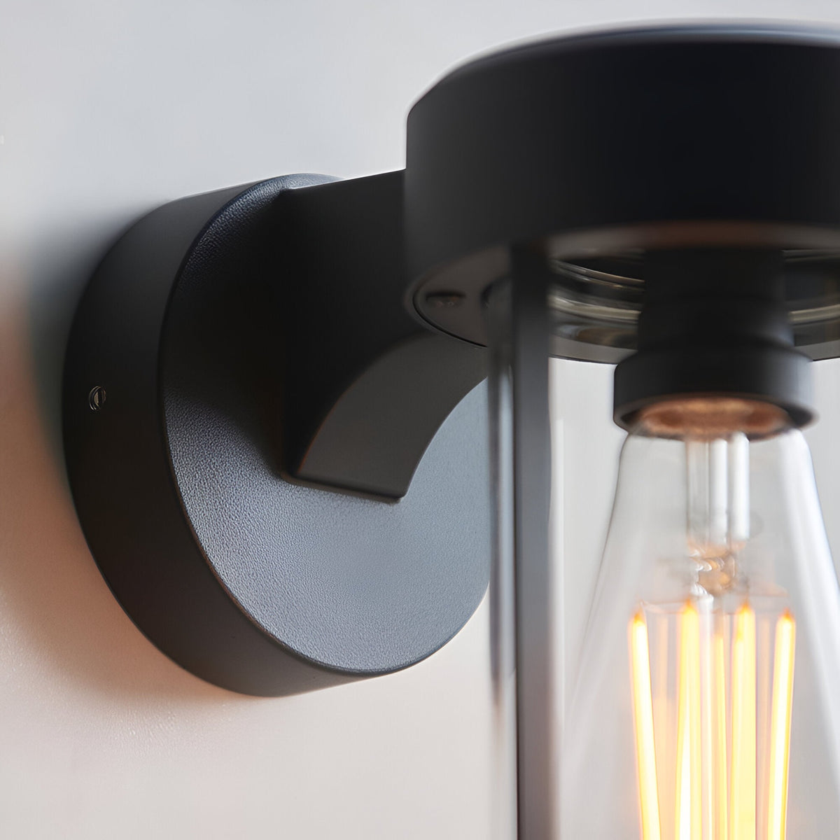 Close-up of the Caldwell Wall Light in matt black die-cast aluminium, showing the round wall mount and clear glass shade with an exposed E27 filament bulb.