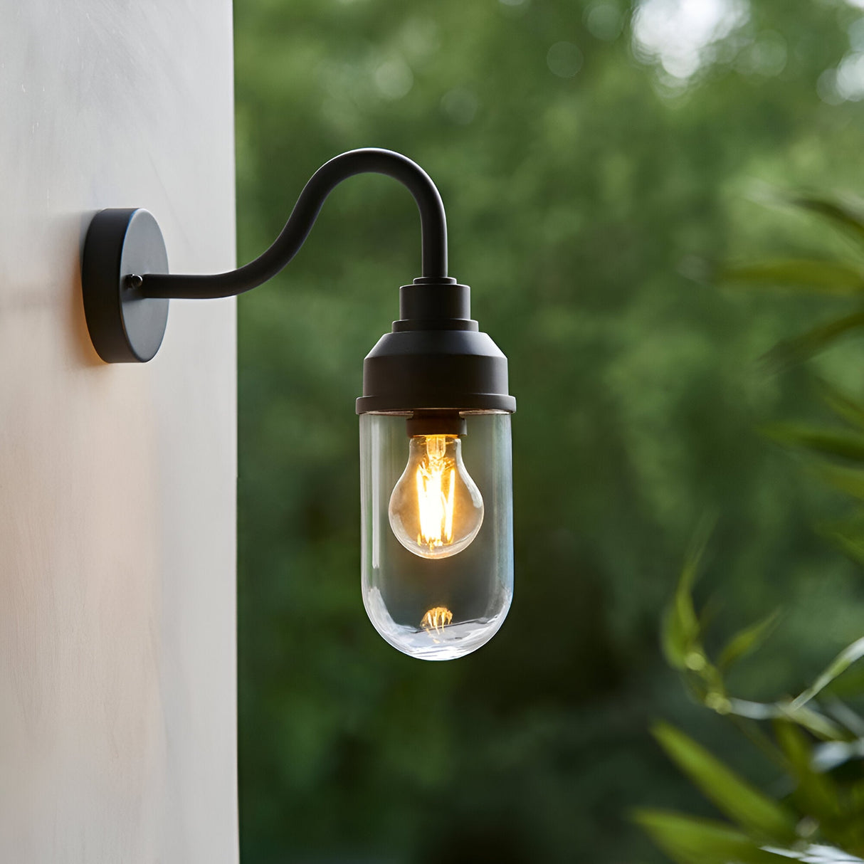 Endon Cranworth matt black wall light with curved gooseneck arm and clear glass shade housing an E27 filament bulb, mounted on a white exterior wall.