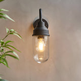 Endon Cranworth matt black wall light with curved arm and clear glass shade housing an E27 filament bulb, mounted on a neutral wall beside foliage.