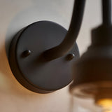 Close-up of the Endon Cranworth wall light's matt black die-cast aluminium base plate and curved arm, with the clear glass shade softly lit alongside.
