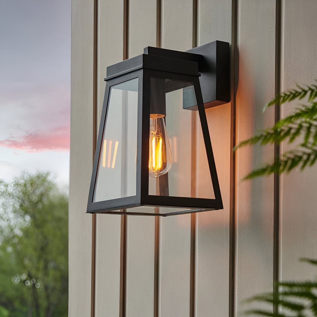 Caldwick Wall Light in matt black, a lantern-style fixture with clear glass panels and exposed bulb, mounted outdoors on light-coloured panelled siding.