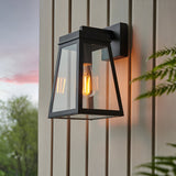 Caldwick Wall Light in matt black, a lantern-style fixture with clear glass panels and exposed bulb, mounted outdoors on light-coloured panelled siding.