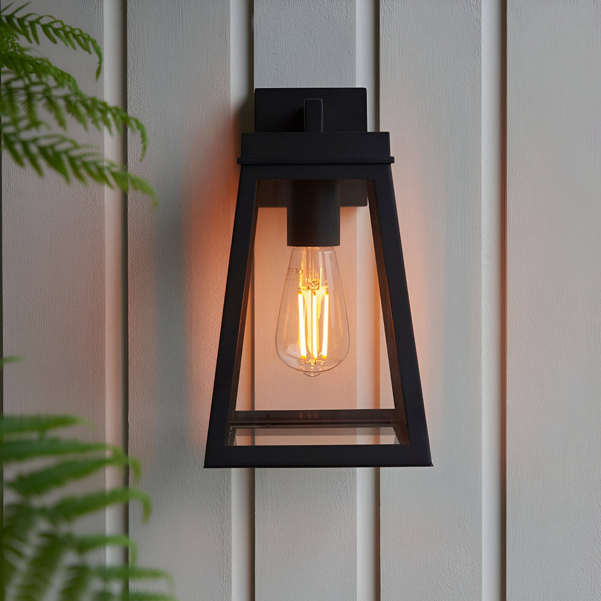 Caldwick Wall Light in matt black, a tapered lantern-style fixture with clear glass panels, mounted on a light wood-panelled wall beside greenery.