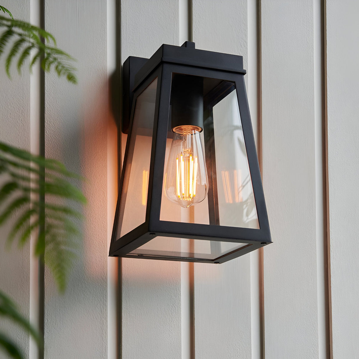 Caldwick Wall Light in matt black, a lantern-style fixture with clear glass panels and visible E27 bulb, mounted on light grey exterior cladding.