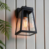 Caldwick Wall Light in matt black, a lantern-style fixture with clear glass panels and visible E27 bulb, mounted on light grey exterior cladding.