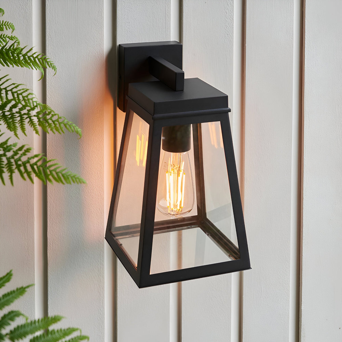 Caldwick Wall Light in matt black, a lantern-style fixture with clear glass panels, mounted on light wood-panelled exterior wall with fern leaves in view.