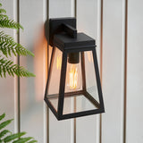 Caldwick Wall Light in matt black, a lantern-style fixture with clear glass panels, mounted on light wood-panelled exterior wall with fern leaves in view.