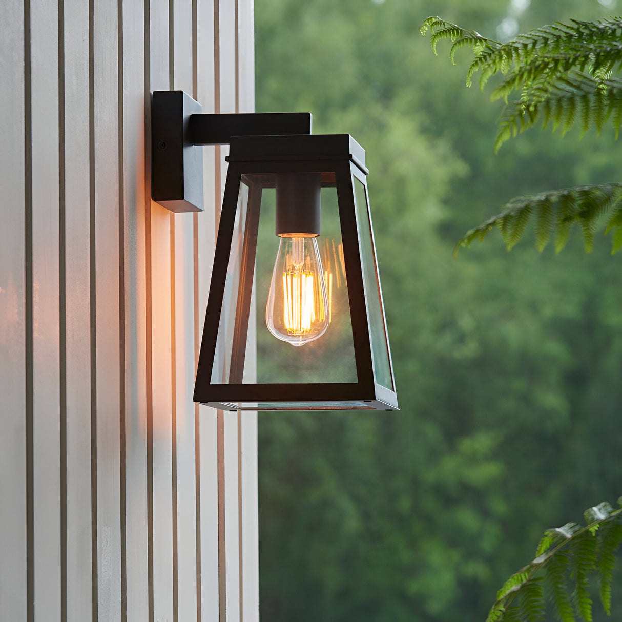 Caldwick Wall Light in matt black, a lantern-style fixture with clear glass panels and exposed bulb, mounted on exterior wall cladding.