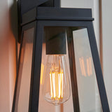 Caldwick Wall Light in matt black, close-up of the lantern-style frame with clear glass panels and a visible E27 filament bulb inside.