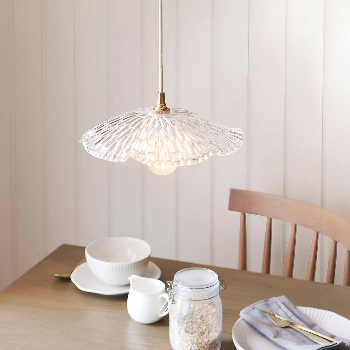 Endon Castshaw pendant light with rippled clear glass shell-shaped shade and brass fitting, hanging above a wooden dining table set with bowls and a jar.