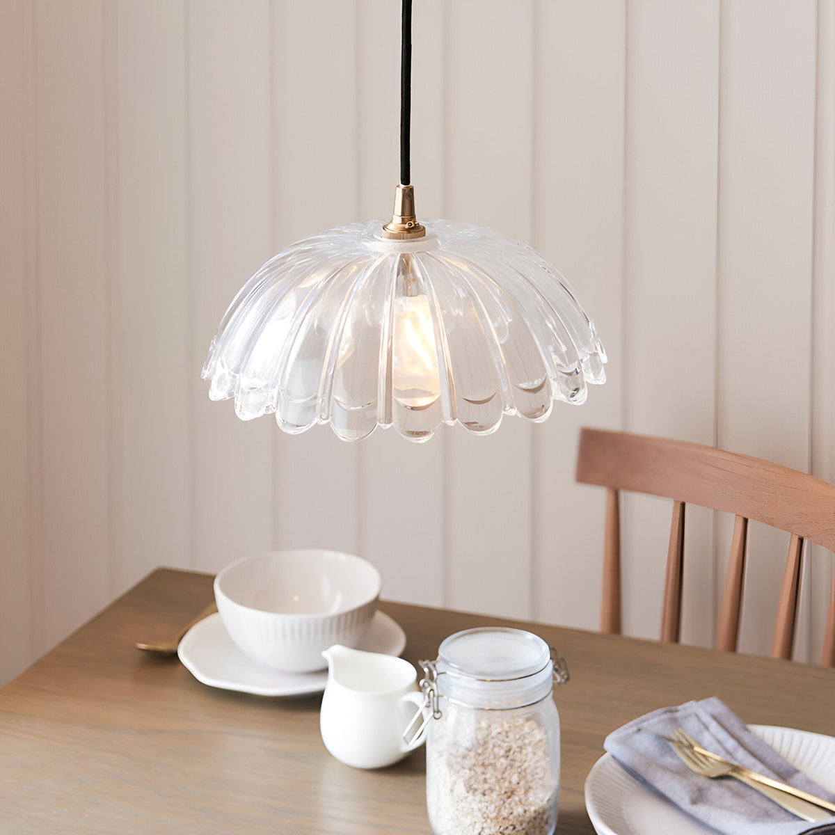Endon Bevcombe scalloped clear glass domed pendant shade with brass fitting on black flex, hung above a wooden dining table set with bowl and jar.
