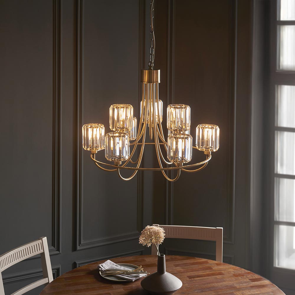Clementine 9 Light Tiered Chandelier in antique brass with faceted crystal glass shades, shown suspended above a dining table in a dark panelled room.