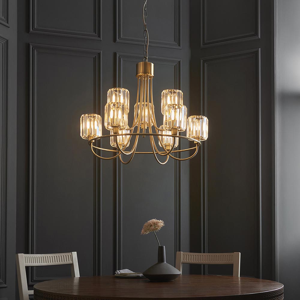 Clementine 9 Light Tiered Chandelier in antique brass with faceted crystal glass shades, hung above a dining table against dark panelled walls.
