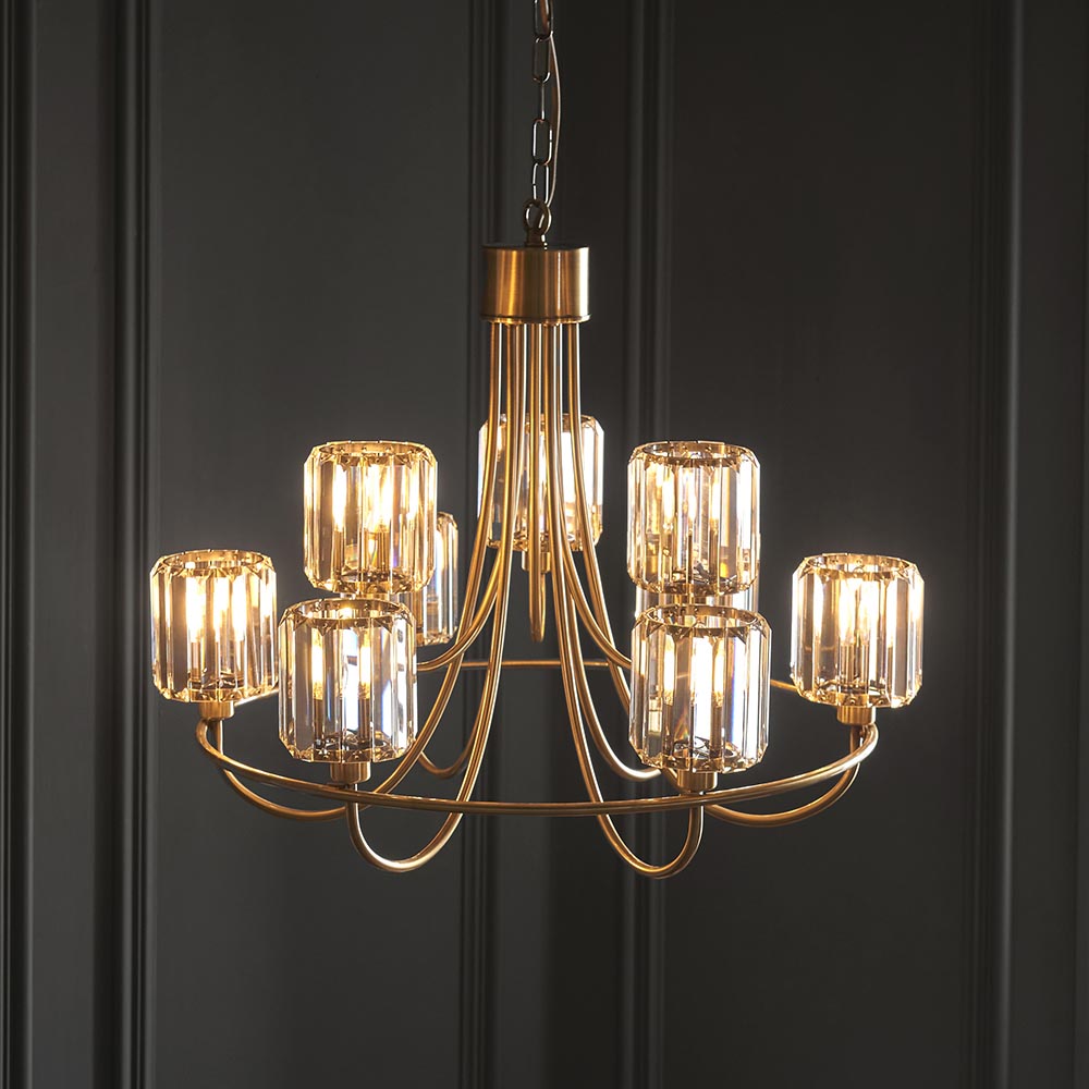 Clementine 9 Light Tiered Chandelier in antique brass with faceted crystal glass shades, hung on a chain against a dark panelled wall.