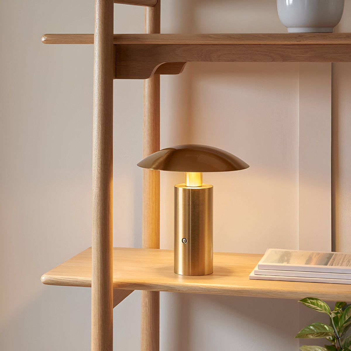 Cranbourne LED Table Lamp in brushed brass with mushroom-dome shade, lit warm white on a wooden shelf beside books and a ladder-style unit.