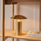 Cranbourne LED Table Lamp in brushed brass with mushroom dome shade, glowing warm white on a wooden shelf beside books.