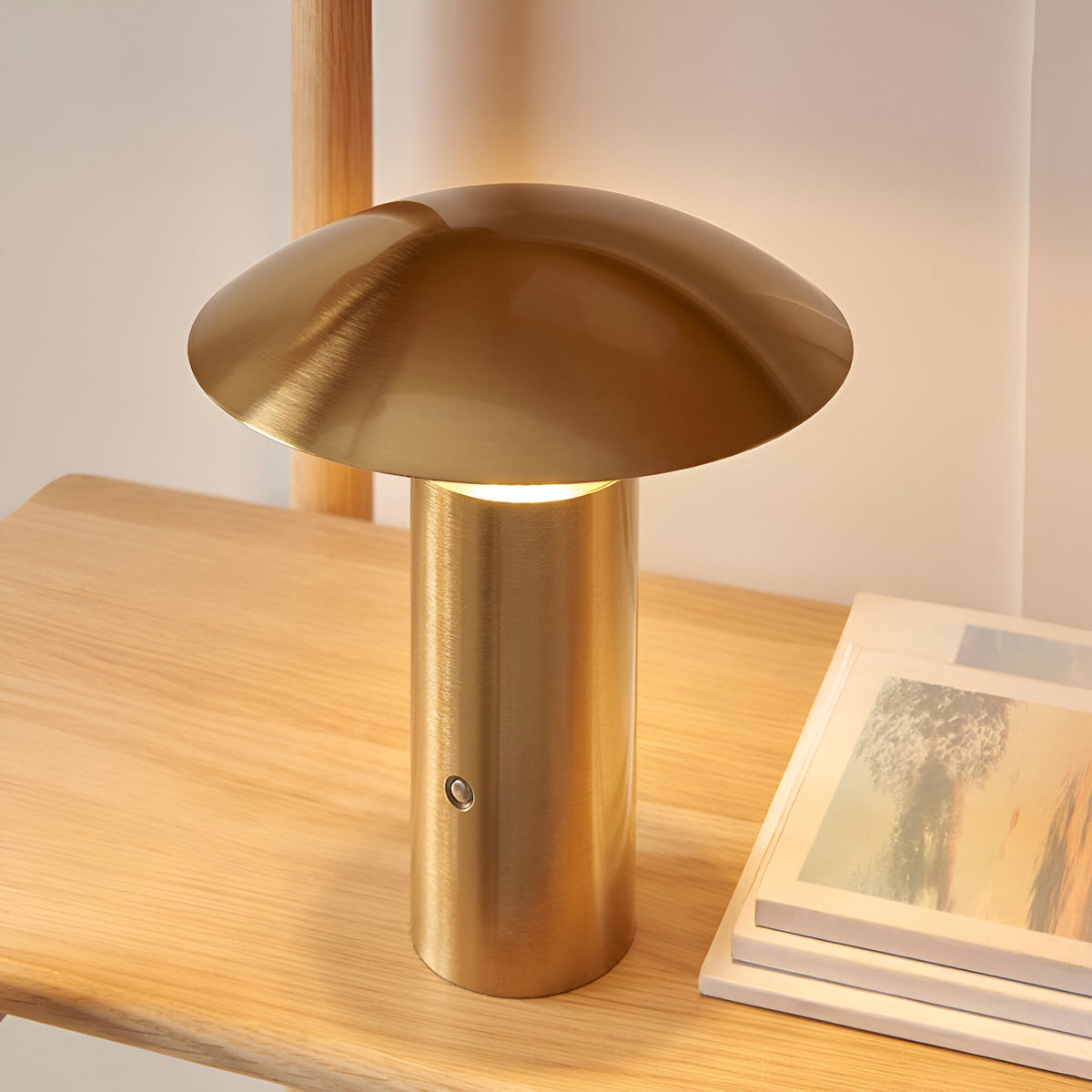 Cranbourne LED Table Lamp in brushed brass with mushroom-dome shade and touch dimmer button, shown lit on a wooden shelf beside books.