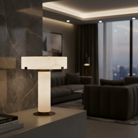 The Virex 1 Light Tiered Column Alabaster Table Lamp – Soft White emits a gentle glow in a chic living room with dark sofas, marble walls, and floor-to-ceiling windows that reveal a stunning city view at dusk.