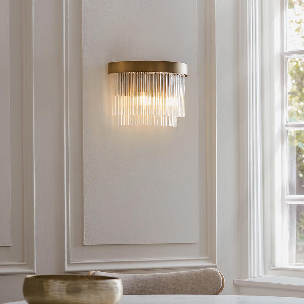 Chatsworth 2 Light Wall Light in antique brass with cascading clear glass rods, illuminated on a panelled wall in a living room setting.