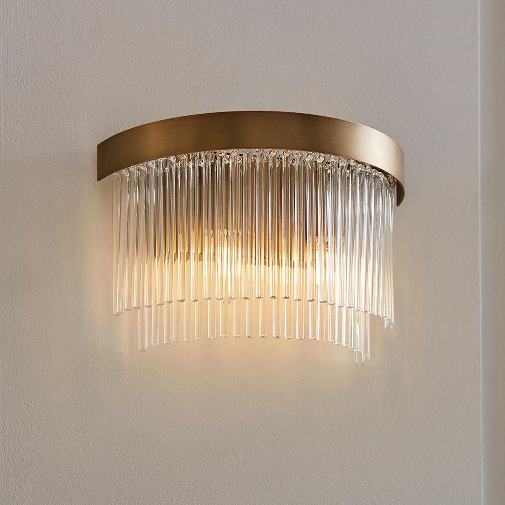 Chatsworth 2 Light Wall Light in antique brass, with curved metal backplate and cascading clear glass rods, mounted on a light wall and softly illuminated.