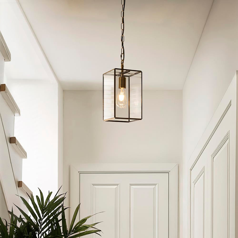 Haddon Pendant in brushed brass, a lantern-shaped fixture with clear glass panels and exposed bulb, hung on chain in a hallway with white doors.