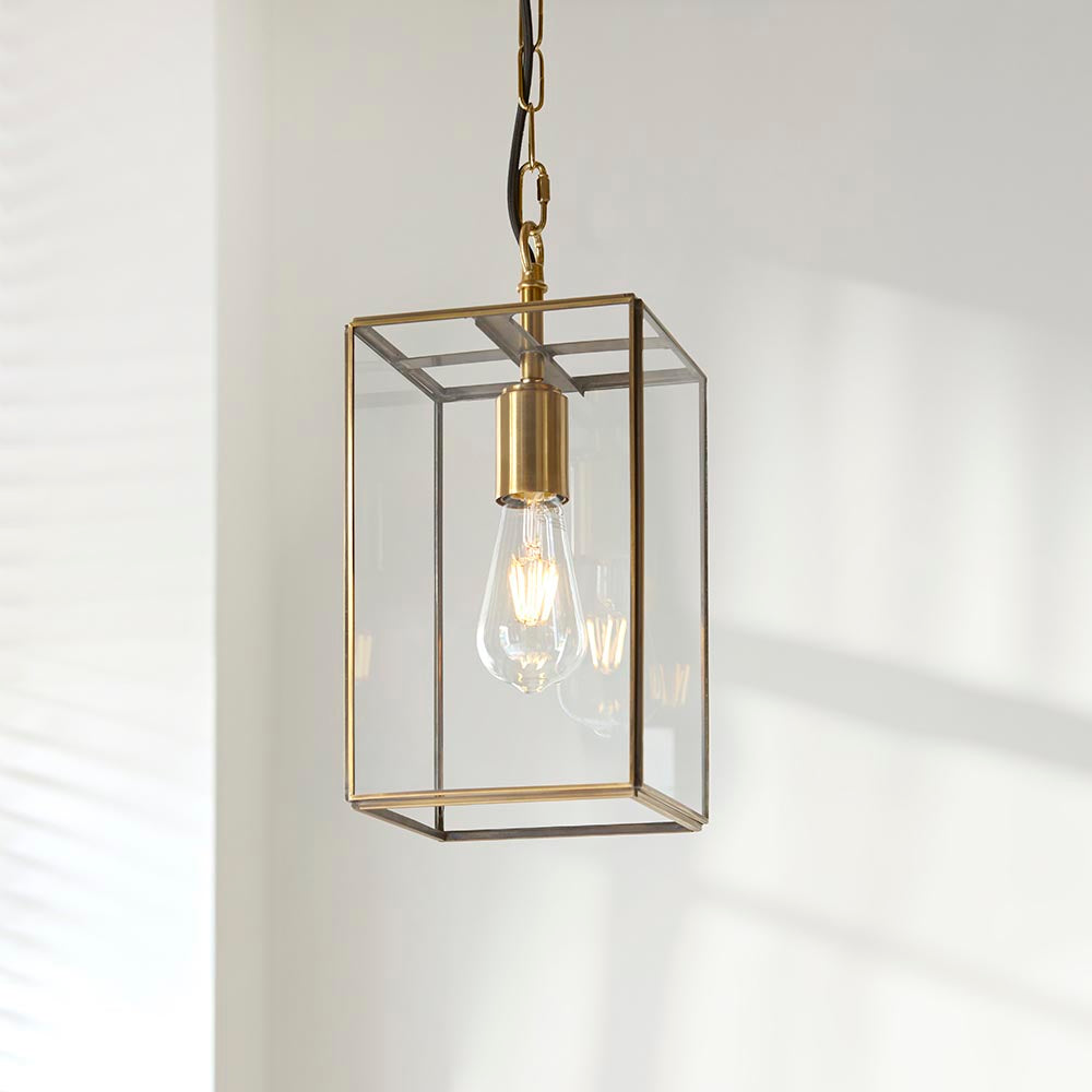 Haddon Pendant in brushed brass with a square lantern frame, clear glass panels, exposed E27 filament bulb, hung from a chain against a plain light wall.