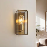 Haddon Wall Light in antique brass, a box-lantern fixture with clear glass panels and exposed filament bulb, shown mounted on a wall in a living room setting.