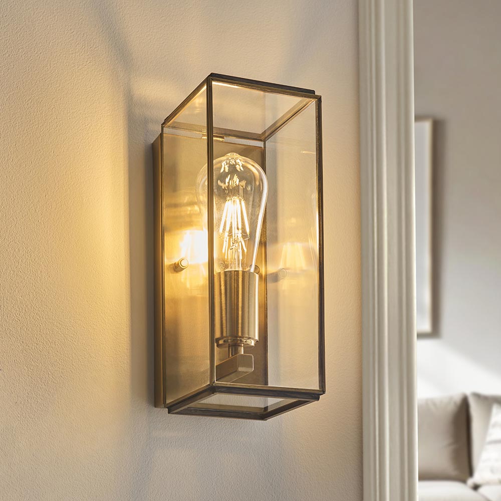 Haddon Wall Light in antique brass, a box-lantern fixture with clear glass panels and exposed filament bulb, shown illuminated on an interior wall.