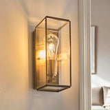 Haddon Wall Light in antique brass, a box-lantern fixture with clear glass panels and exposed filament bulb, shown illuminated on an interior wall.