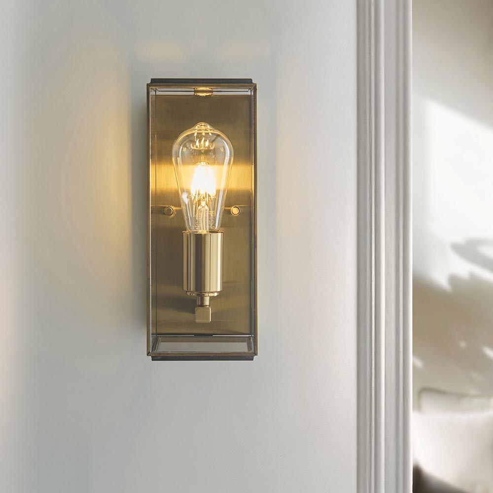 Haddon Wall Light in antique brushed brass, a box-lantern fixture with clear glass panels and exposed filament bulb, mounted on a light grey wall.