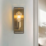 Haddon Wall Light in antique brushed brass, a box-lantern fixture with clear glass panels and exposed filament bulb, mounted on a light grey wall.