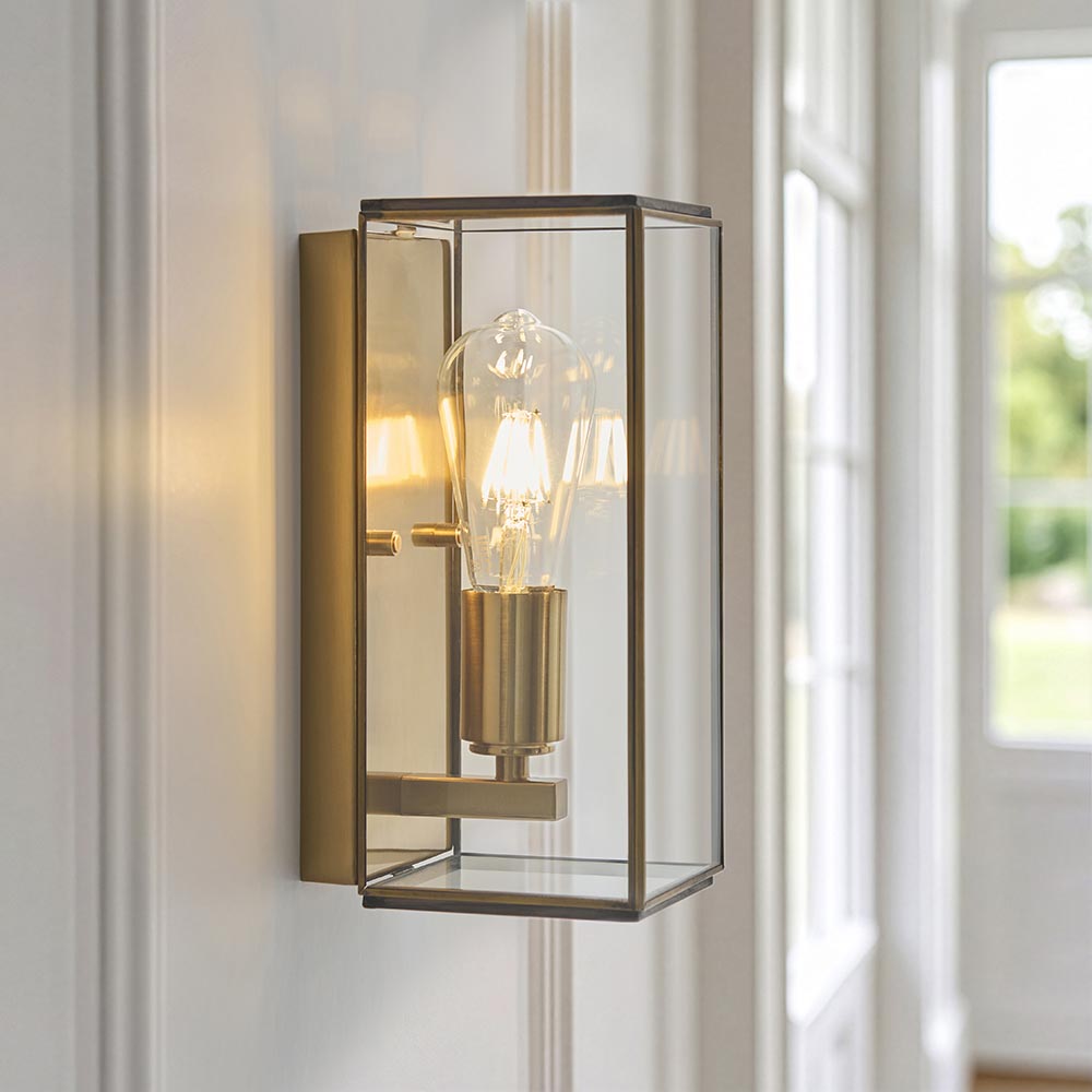Haddon Wall Light in antique brass, a box-lantern design with clear glass panels and visible filament bulb, mounted on a hallway wall.
