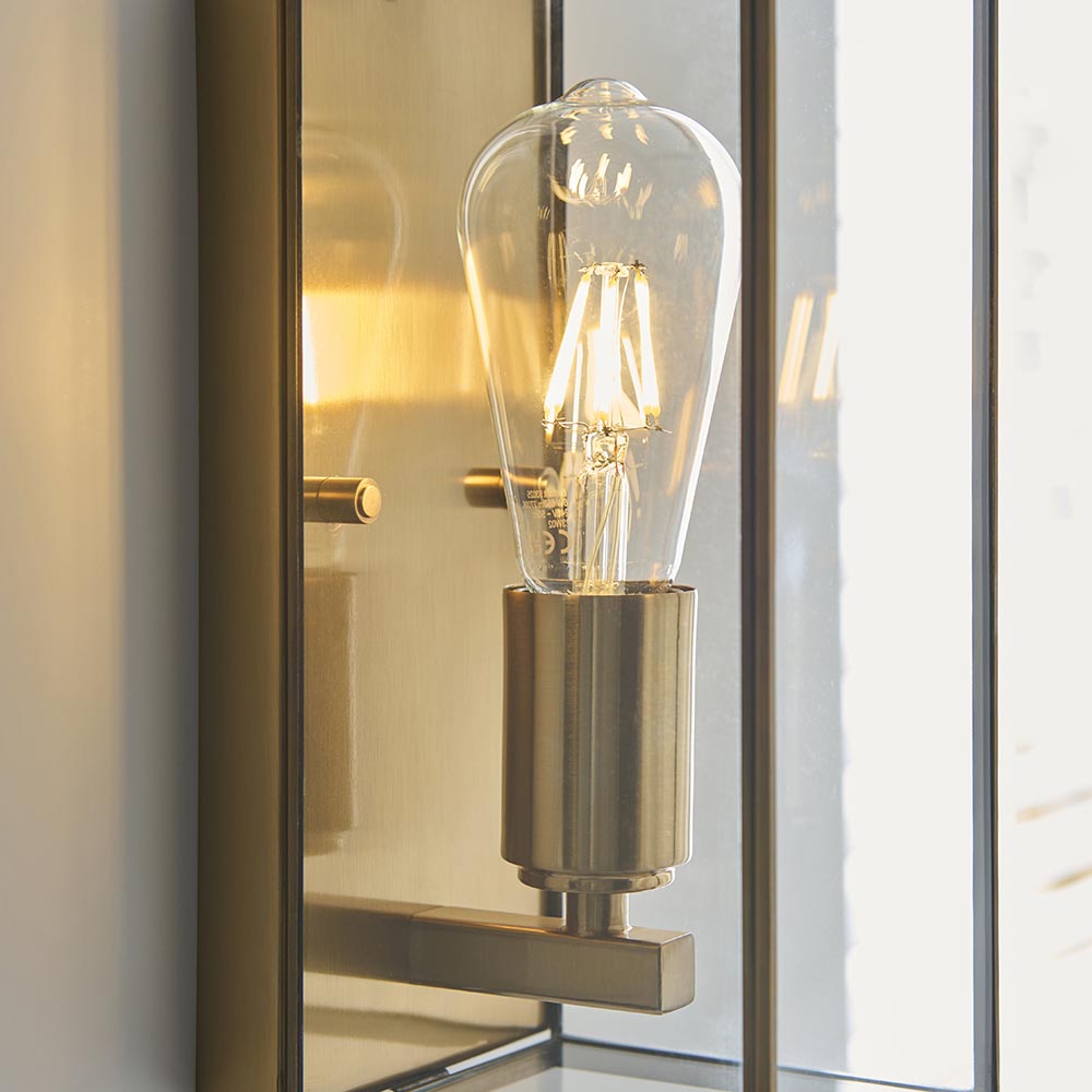 Close-up of the Haddon Wall Light in antique brass, box-lantern frame with clear glass panels showing an E27 filament bulb on a brushed brass holder.