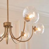 Endon Bramwick 7 Light Pendant in antique brass with curved arms and clear twisted glass globes, shown lit against a panelled cream wall.