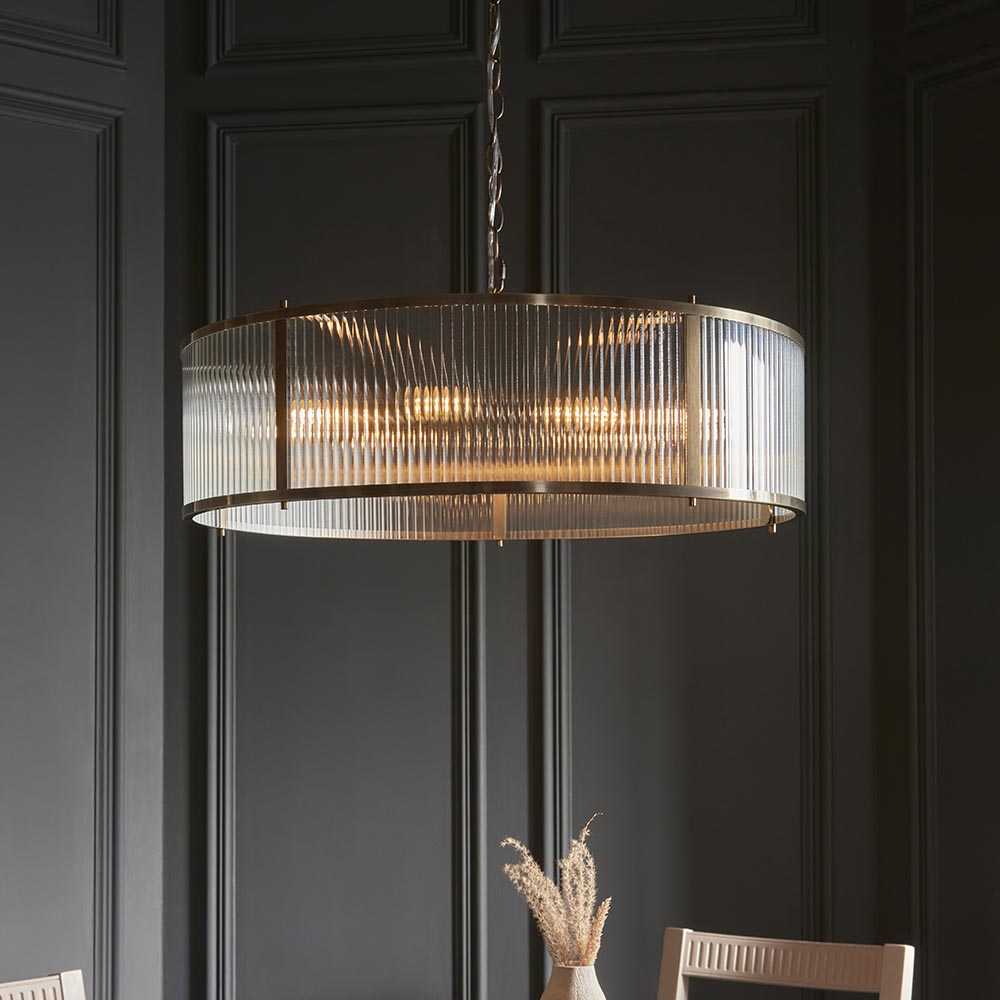 Endon Rothbury 8 light pendant in antique brass with a drum frame of clear ribbed glass panels, lit above a dining setting against dark panelled walls.