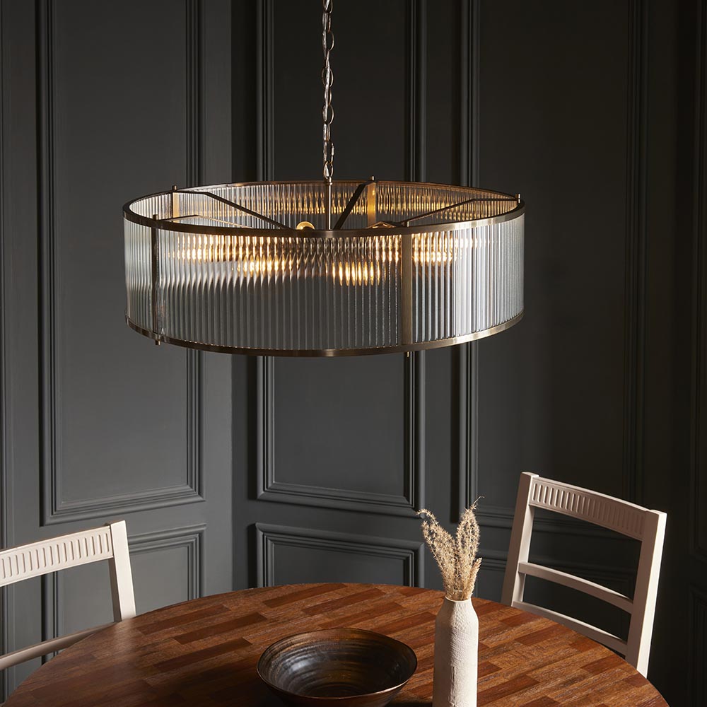 Endon Rothbury 8 light pendant in antique brass with clear ribbed glass drum shade, hung above a round wooden dining table against dark panelled walls.