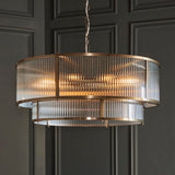 Endon Rothbury 8 light tiered chandelier in antique brass with clear ribbed glass panels over two tiers, suspended from a chain against a grey panelled wall.