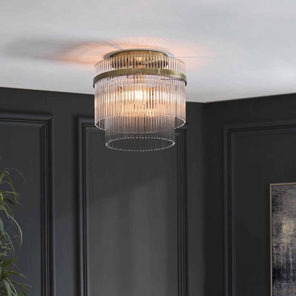Endon Beaumont flush ceiling light in antique brass with tiered glass rod shade, illuminated on a white ceiling above dark panelled walls.