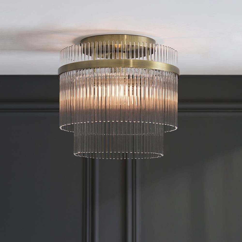 Endon Beaumont two-tier flush ceiling light in antique brass with vertical glass rod shades, lit and mounted to a white ceiling above dark panelled walls.