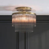 Endon Beaumont two-tier flush ceiling light in antique brass with vertical glass rod shades, lit and mounted to a white ceiling above dark panelled walls.