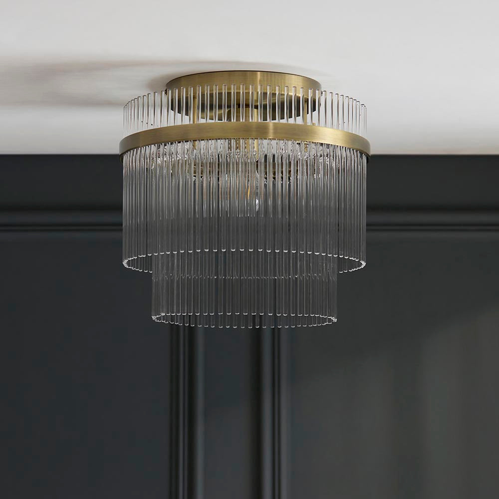 Endon Beaumont flush ceiling light in antique brass, with a two-tier glass rod shade mounted against a white ceiling above a dark panelled wall.