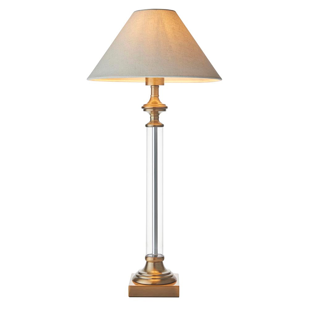 Aldmont Avalon Table Lamp with a clear glass column, antique brass base and cap, and a tapered natural linen shade, shown on a plain white background.