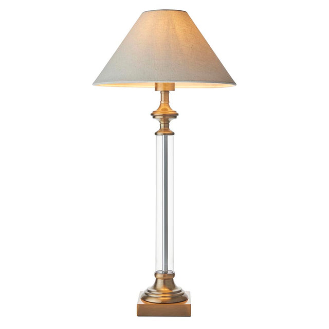 Aldmont Avalon Table Lamp with a clear glass column, antique brass base and cap, and a tapered natural linen shade, shown on a plain white background.